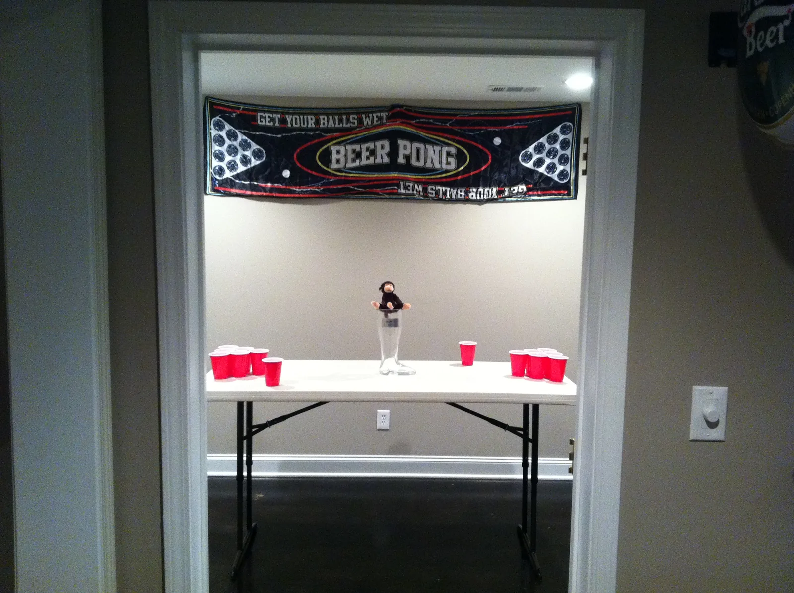 Empty room through a doorway: beer pong table, red cups, a glass boot with a stuffed monkey, and a GET YOUR BALLS WET banner