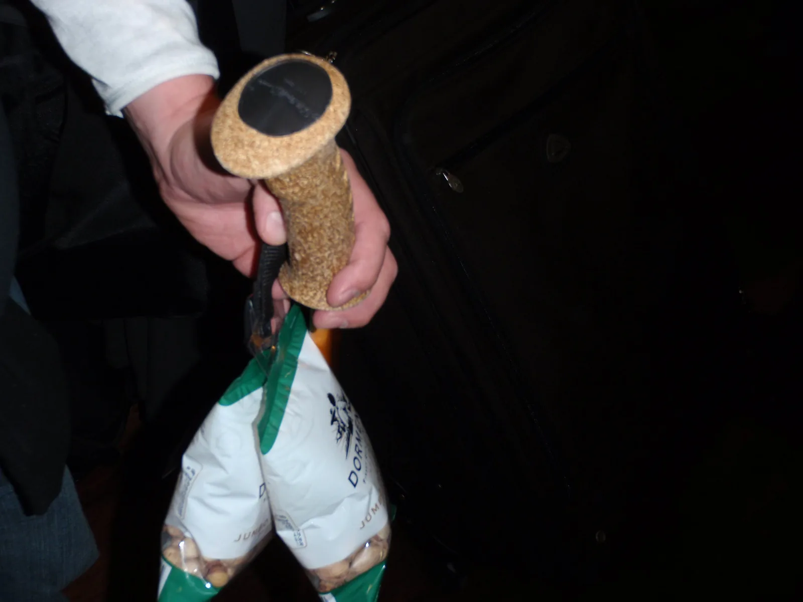 A hand holds a cork cane handle. Two bags of pistachios hang from the handle beside a suitcase.
